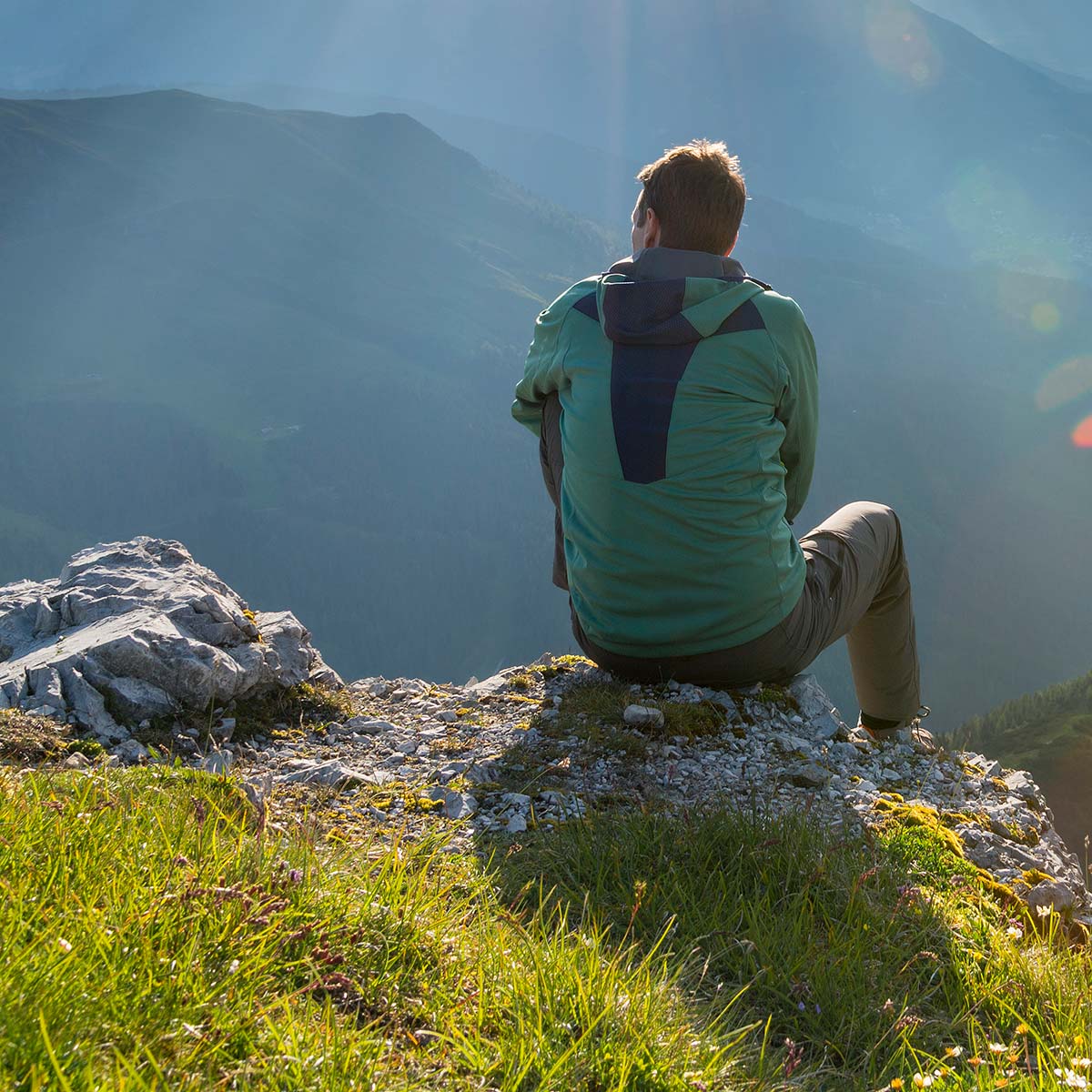 A man sits on a rock, looking over a beautiful green landscape, enjoying a peaceful moment in nature