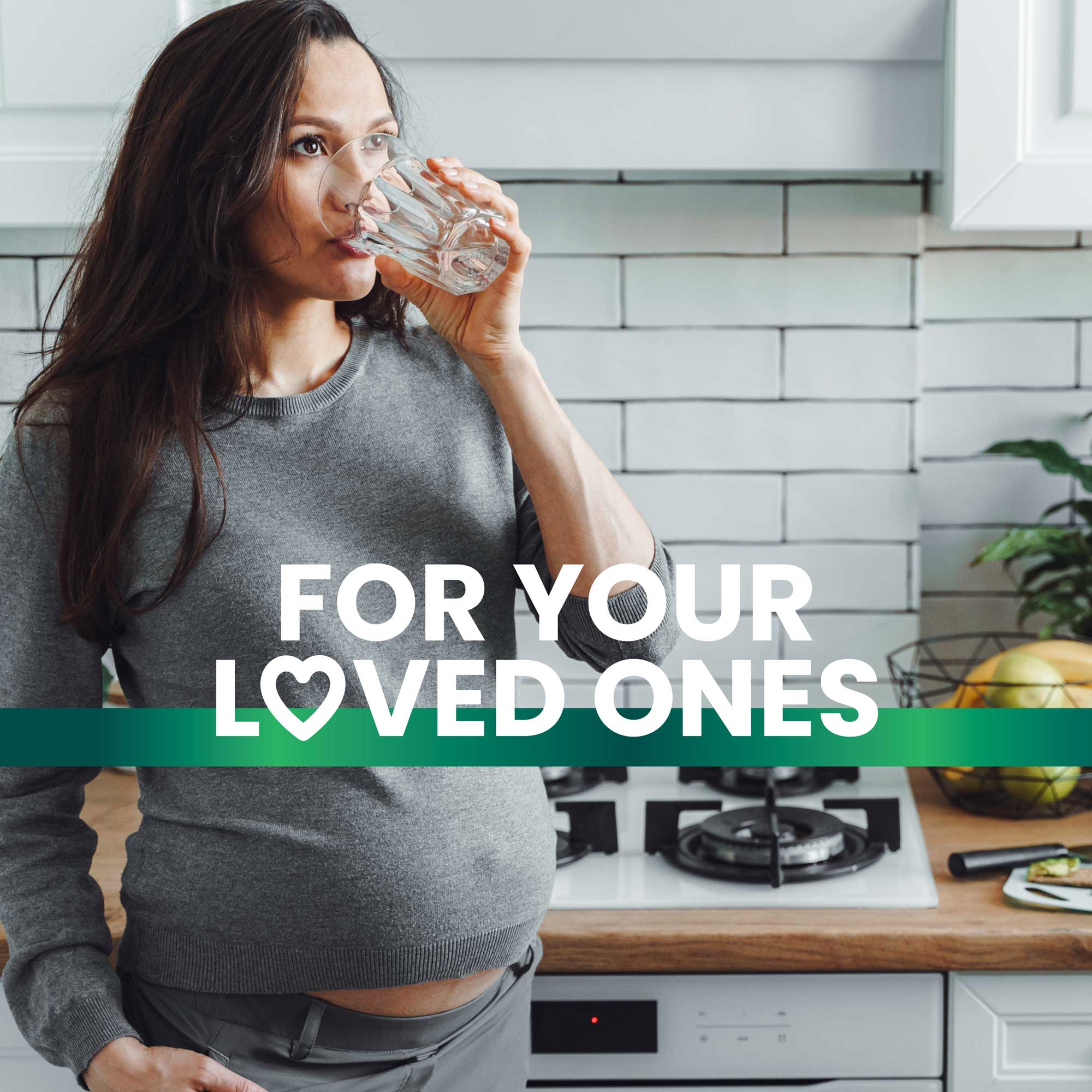 A pregnant woman stands in the kitchen, drinking a glass of water with a smile, highlighting healthy hydration and promoting a happy, healthy lifestyle. Text overlay reads "For your loved ones."